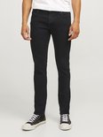 Man met een witte top en zwarte skinny jeans, staand op een lichtgrijze achtergrond.