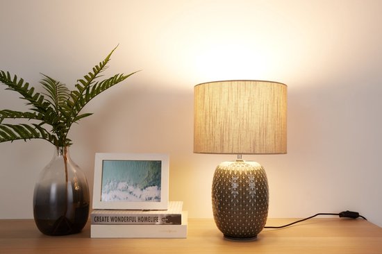 Grijze tafellamp met beige kap en decoratieve voet, verlicht op een houten tafel naast een vaas en boeken.