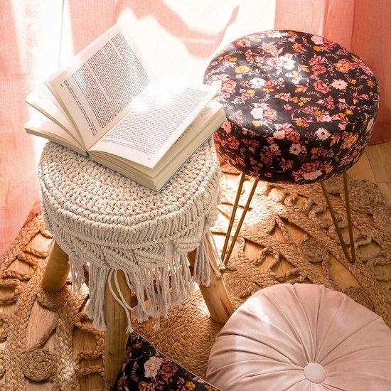 Een beige macramé krukje met een open boek erop, naast een bloemige kruk en een roze kussen.