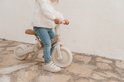 Onderlichaam van een kind in spijkerbroek en witte schoenen zittend op een crèmekleurige loopfiets met bloemenprint.