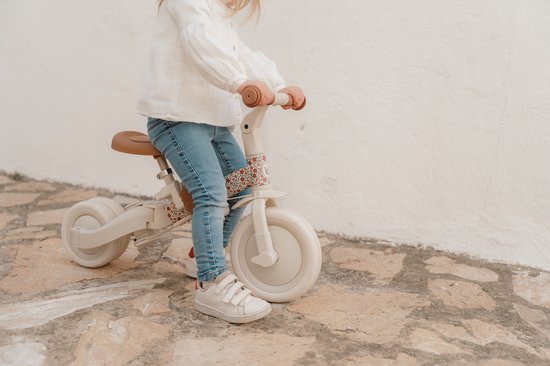 Onderlichaam van een kind in spijkerbroek en witte schoenen zittend op een crèmekleurige loopfiets met bloemenprint.