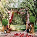 Decoratieve bruiloftsbrug versierd met bloemen, draperieën, lampjes en twee buxusbollen op houten standaarden.