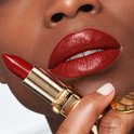 Een vrouw met een donkere huid, volle rode lippen en rode nagels houdt een gouden L'Oréal lipstick vast. De lippenstiftkleur is diep rood.