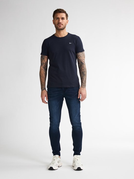 Man met tatoeages draagt een marineblauwe T-shirt en een blauwe skinny jeans met witte sneakers.