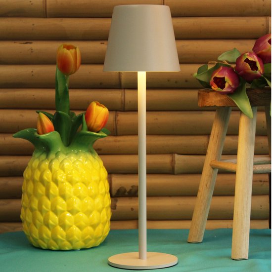 Witte tafellamp met grijze kap naast een gele ananasvaas met tulpen en een deel van een houten kruk met tulpen.
