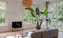 Zwarte Harman Kardon soundbar op een lage tv-standaard, in een woonkamer met een grote plant en grijze bank.