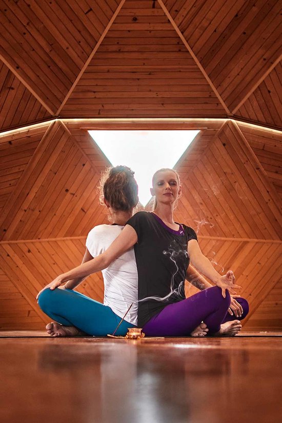 Twee vrouwen in yoga-outfits, zittend met de rug tegen elkaar in een houten geometrische ruimte. Paarse en blauwe legging.