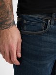 Close-up van de voorkant van een donkerblauwe jeans met oranje stiksels en een tatoeage op de onderarm.