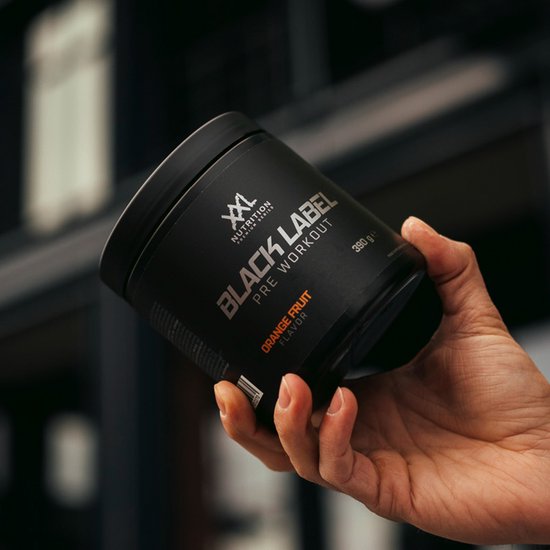 Zwarte pot met 'XXL Nutrition BLACK LABEL PRE WORKOUT ORANGE FRUIT FLAVOR 390 g' in zilveren en oranje letters, vastgehouden door een hand.