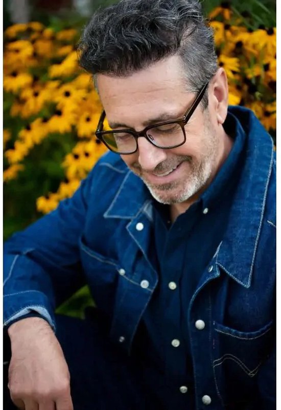 Homme portant des lunettes à monture brun foncé, une veste en jean, souriant, à l'extérieur avec des fleurs jaunes en arrière-plan.