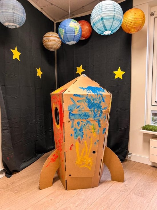 Buitenkant van een kartonnen raket versierd met kinderlijke schilderingen in blauw, rood en geel. Met planeetdecoratie aan het plafond.