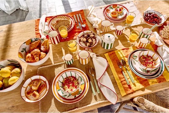 Een gedekte tafel met ontbijt, waaronder fruit, croissants, gebak en citroenen, met servies met strepen en fruitmotieven.
