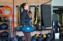 Blauwe JBL Xtreme speaker met schouderband, gedragen door een vrouw in een fitnessruimte.