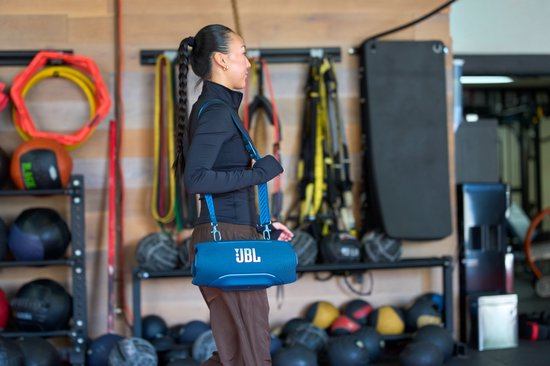 Blauwe JBL Xtreme speaker met schouderband, gedragen door een vrouw in een fitnessruimte.