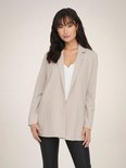 Vrouw met lange, golvende donkere haren in een lange, beige blazer met open voorkant en een witte top.