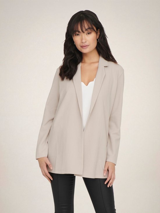 Vrouw met lange, golvende donkere haren in een lange, beige blazer met open voorkant en een witte top.