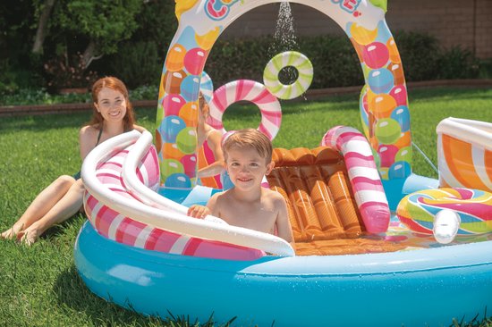 Een jongen in het 'Candy Zone' speelbad met glijbaan, met een glimlachende vrouw op de achtergrond.