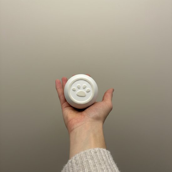 Een hand houdt een kleine ronde witte urn omhoog met een pootafdruk reliëf op de bodem.