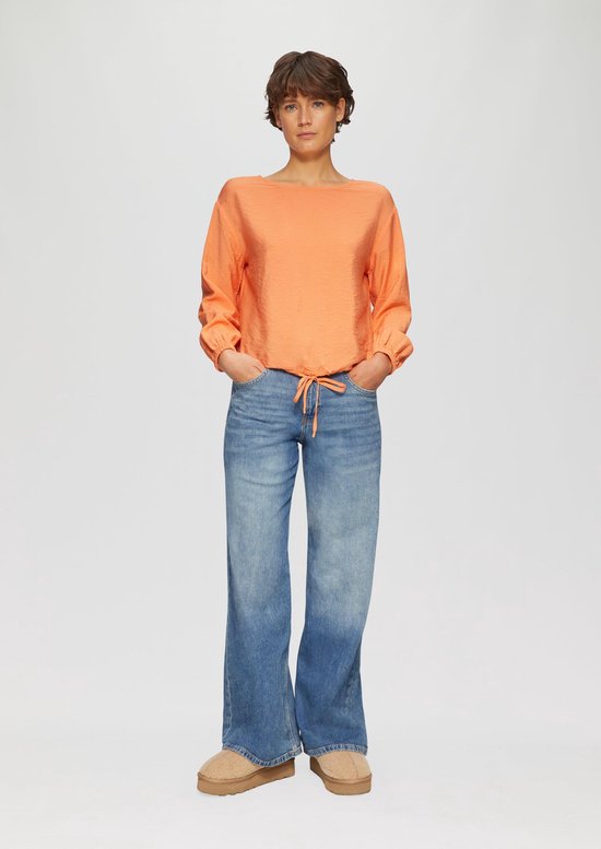 Model draagt oranje s.Oliver blouse en blauwe jeans, met handen in zakken.