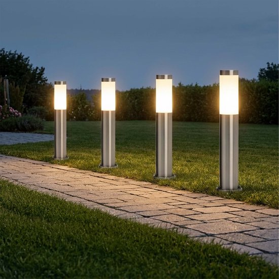Vier zilverkleurige tuinlampen langs een tuinpad, 's avonds brandend, op gras.