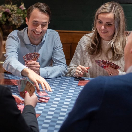 Vier mensen spelen een kaartspel aan tafel met een blauw-wit geblokt tafelkleed. Ze houden kaarten vast en lachen.