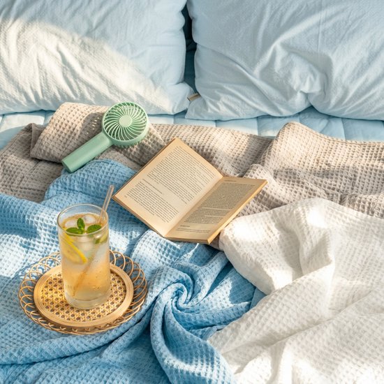 Een open boek, een glas ijsthee en een ventilator liggen op blauwe, grijze en witte wafelstof dekens.