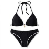 Zwarte triangelbikini met witte zigzagrand en schelpbedeltje, plat gelegd op witte achtergrond.