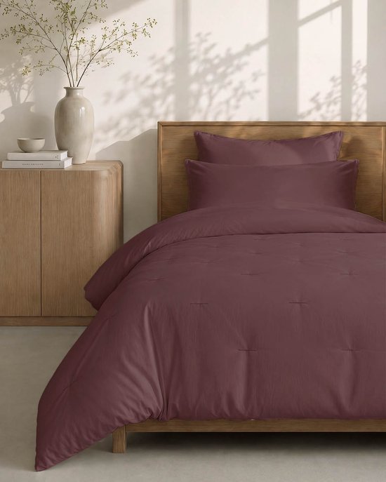Een opgemaakt bed met een bordeauxrood dekbed en kussenslopen, rustend op een houten bedframe.