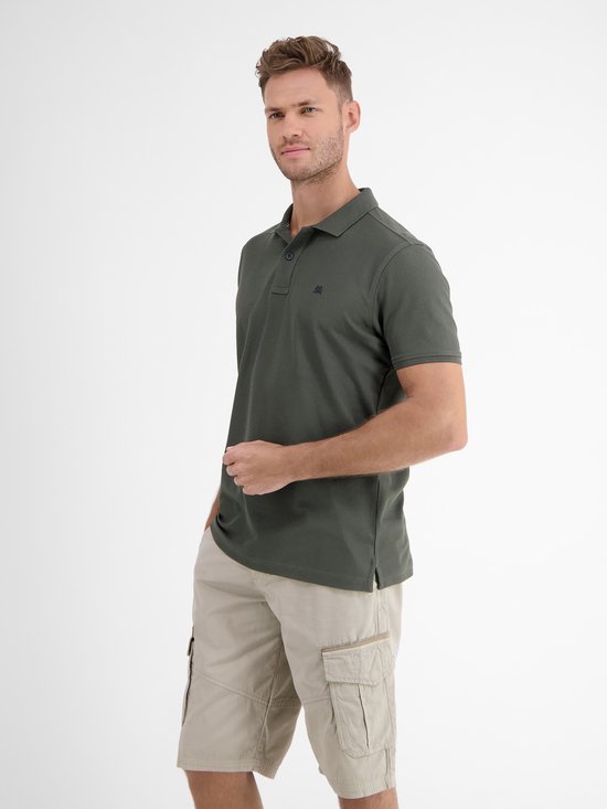 Man met donkergroen poloshirt en beige cargo shorts, staand met een hand in de zak.