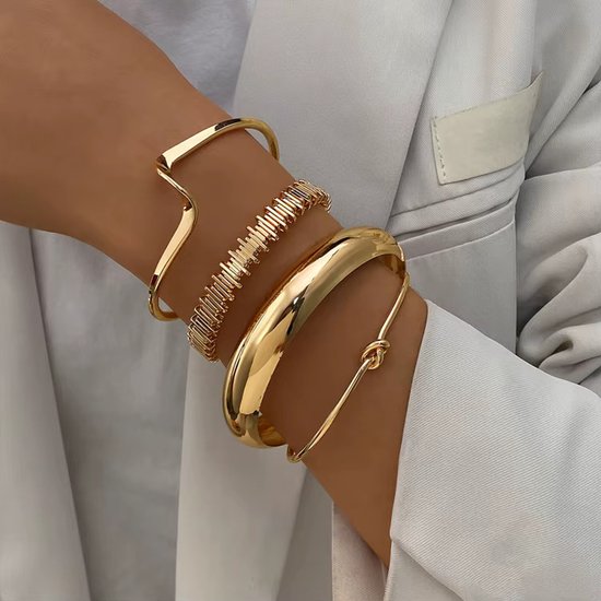 Een set van vier gouden armbanden gedragen op een damespols. De set bevat een brede gladde bangle, een golvende cuff, een textuur armband en een dunne armband met knoop.