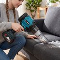 Een vrouw met grijze trui en spijkerbroek gebruikt een blauwe/zwarte stofzuiger om pluizen van een grijze bank te zuigen.
