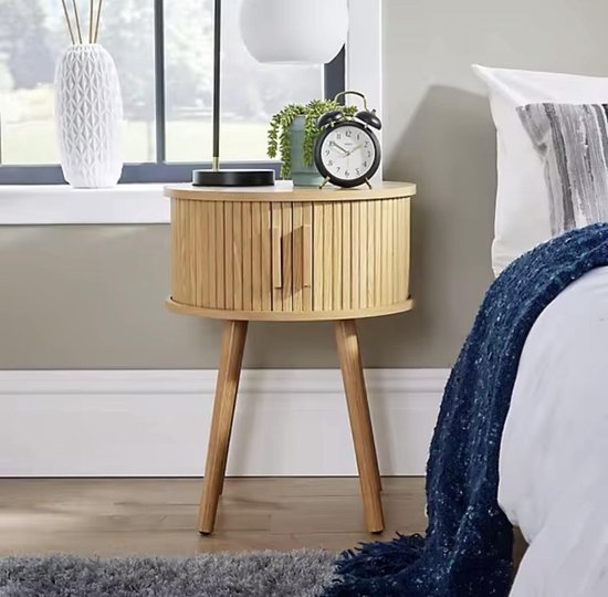 Ronde houten bijzettafel met geribbelde zijkanten en een deurtje, naast een bed met een donkerblauwe deken. Een wekker staat op de tafel.