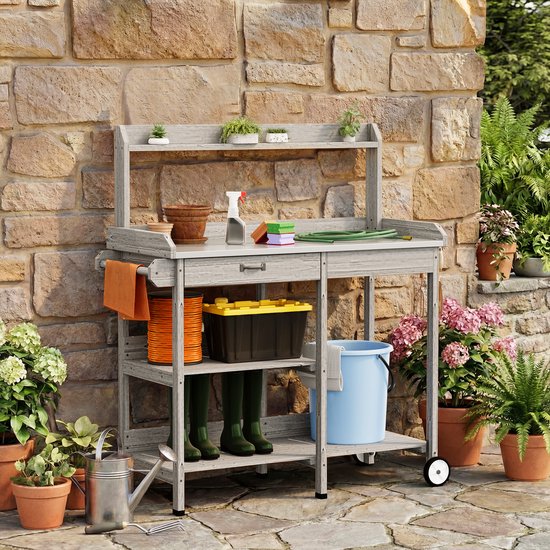 Grijze houten plantentafel met plantenbakken, een sproeier, tuinslangen, groene laarzen en een blauwe emmer.