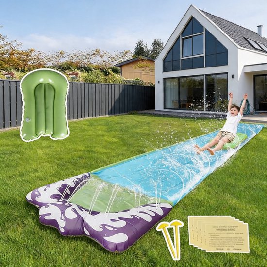 Jongen glijdt op de paarse, witte en groene waterglijbaan in de achtertuin, met een opblaasbaar groen kussen.