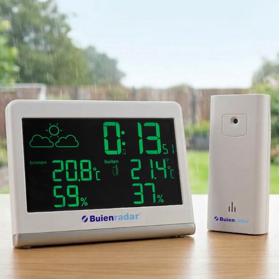 Een wit digitaal weerstation en een witte buitensensor, beide met groen display, staan op een houten tafel. Buiten is het regenachtig.