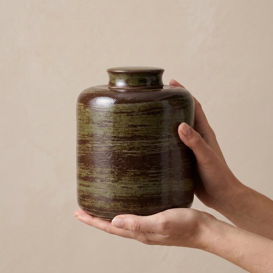 Handen houden een groene en bruine keramische urn vast.