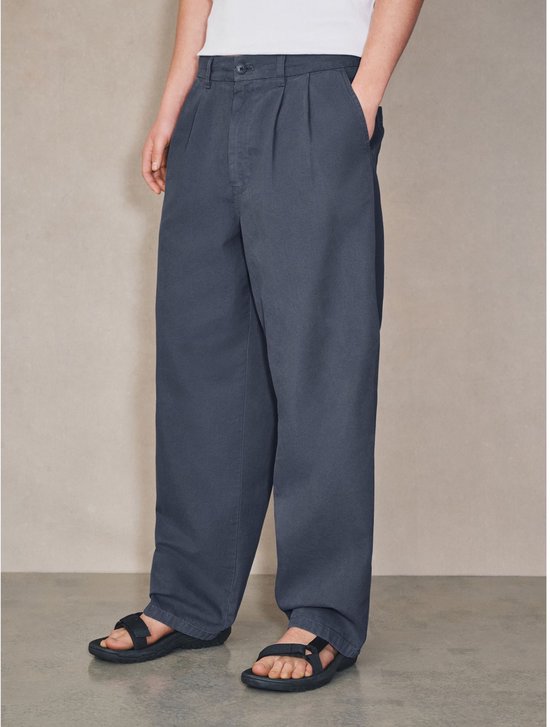 Model draagt een wijde, donkerblauwe broek met dubbele plooien, een knoopsluiting en zwarte sandalen.