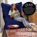 Een vrouw met een lichte trui en spijkerbroek zit op een grote, donkerblauwe zitzak op een balkon. Ze houdt een kopje vast.