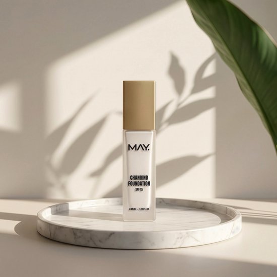 Gouden pompflacon "CHANGING FOUNDATION SPF 15" op marmeren plateau met plantenbladeren op achtergrond.