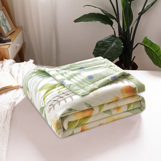 Een opgevouwen lichtgroen dekbed met een bloemenprint van gele en oranje bloemen en groene bladeren op een bed. Een open boek ligt ernaast.