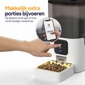 Hand duwt op de voerknop van een witte voerautomaat. Een smartphone toont de "Feed Now" knop. Brokken vallen in de gouden voerbak.