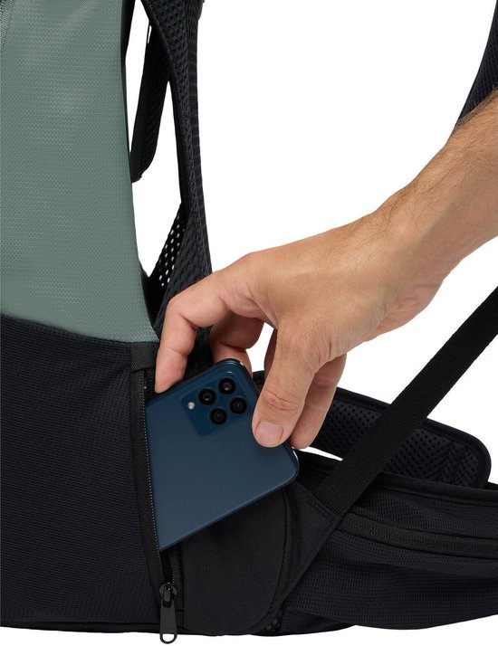 Hand plaatst een blauwe smartphone in een zijvak van de groene rugzak.