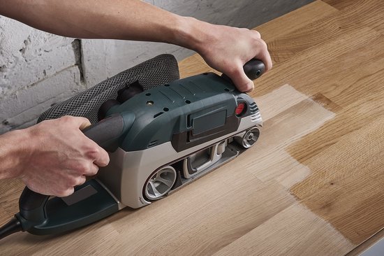 Groene schuurmachine met één band wordt gebruikt op een houten tafel, schuurdeeltjes vliegen rondom.