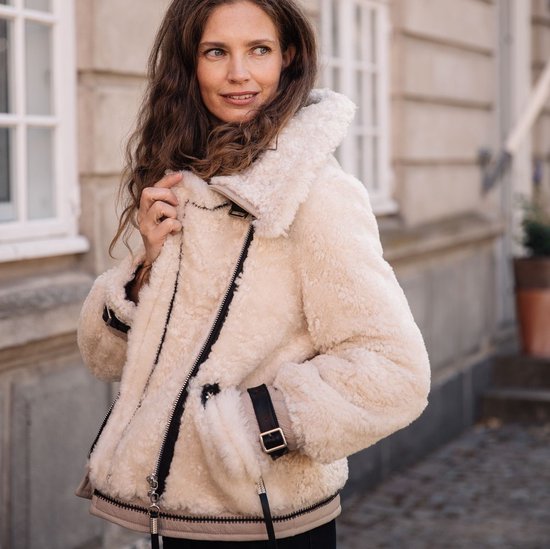 Femme aux longues boucles brunes, porte une veste en shearling crème avec des sangles et des boucles en cuir noir au col.