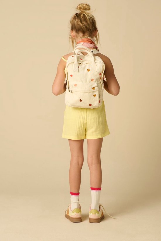 Meisje met een crèmekleurige rugzak met hartjesprint, gele playsuit en roze sokken.