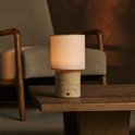 Tafellamp met beige linnenkap en cilindrische travertijnen voet, staand op een houten tafel.