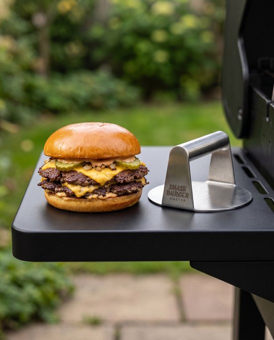 Dubbele smash burger met gesmolten kaas, sla, augurken en saus, naast een RVS pers op een grill.