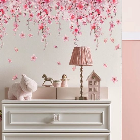 Hangende roze bloesemtakken boven een dressoir met decoraties en een lamp, op een muur met losse bloesems.