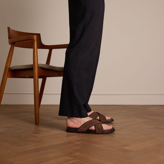 Mannequin portant des mules en daim marron à brides croisées, à côté d'une chaise.