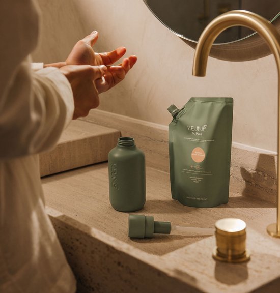 Personne se lavant les mains au-dessus d'un lavabo, à côté d'une bouteille et d'une recharge Keune vertes, robinet doré.